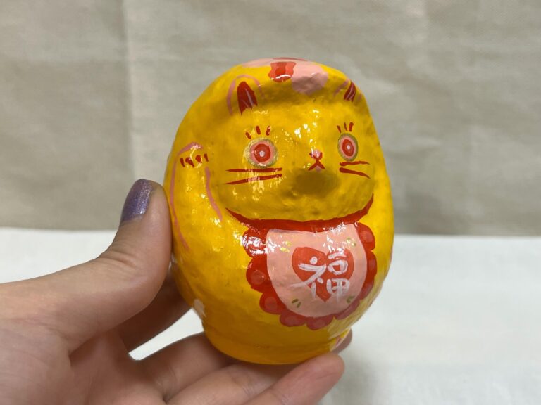 A hand holding a yellow cat Daruma art piece by Kotori/KTR