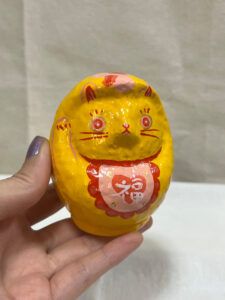 A hand holding a yellow cat Daruma art piece with the character fuku